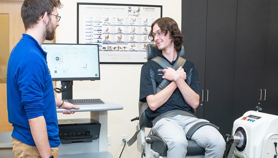 一名参加运动训练的学生坐在一台测试腿部力量的机器上，而另一名学生则在电脑显示器上查看结果
